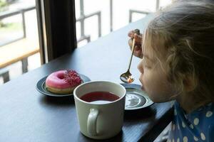 un pequeño niña bebidas té con un rosquilla en un cafetería. acogedor Mañana con dulce comida foto