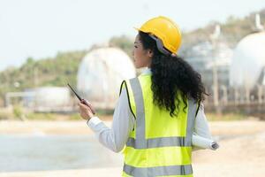 female engineer with hardhat with petrochemical factory background. asian woman holding tablet, plan and Walkie Talkie. photo