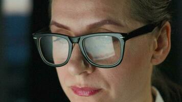 kvinna i glasögon ser på de övervaka och och arbetssätt med diagram och analyser. de övervaka skärm är reflekterad i de glasögon. arbete på natt. Hem kontor. avlägsen arbete video