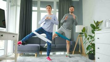 Caucasian couple is doing squats and leg swings with elastic band at home in cozy bright room, slow motion video