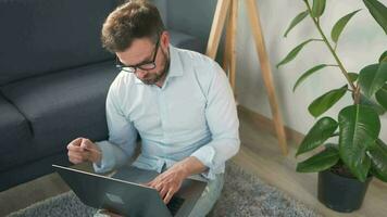 bärtig Mann mit Brille Sitzung auf Teppich mit Laptop und Arbeiten im gemütlich Zimmer. Fernbedienung Arbeit draußen das Büro. video