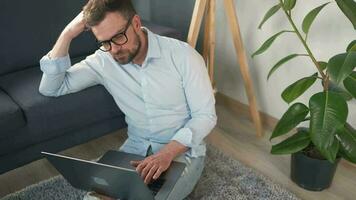 barbu homme avec des lunettes séance sur tapis avec portable et travail dans confortable chambre. éloigné travail à l'extérieur le bureau. video