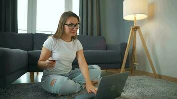 Woman with glasses is sitting on the carpet and makes an online purchase using a credit card and smartphone. Online shopping, lifestyle technology video