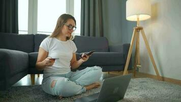 Woman with glasses is sitting on the carpet and makes an online purchase using a credit card and smartphone. Online shopping, lifestyle technology video