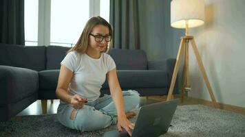 Woman with glasses is sitting on the carpet and makes an online purchase using a credit card and smartphone. Online shopping, lifestyle technology video