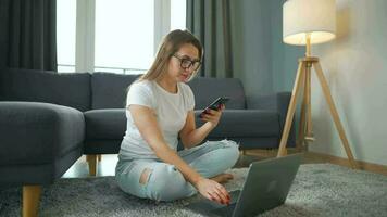 casualmente vestito donna con bicchieri seduta su tappeto con il computer portatile e smartphone e Lavorando nel accogliente camera. lei comunica e lavori a distanza. a distanza opera al di fuori il ufficio video