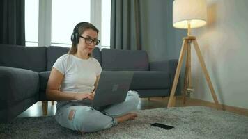 casualmente vestido mulher com fones de ouvido é sentado em tapete com computador portátil dentro acolhedor sala. ela comunica e trabalho remotamente. controlo remoto trabalhos lado de fora a escritório. video