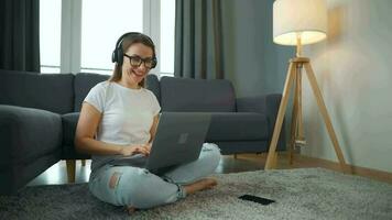 terloops gekleed vrouw met hoofdtelefoons is zittend Aan tapijt met laptop in knus kamer. ze communiceert en werken van een afstand. afgelegen werk buiten de kantoor. video