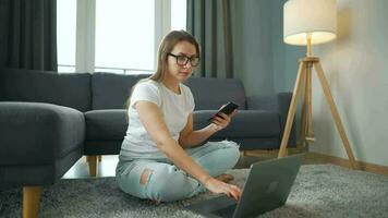 casualmente vestito donna con bicchieri seduta su tappeto con il computer portatile e smartphone e Lavorando nel accogliente camera. lei comunica e lavori a distanza. a distanza opera al di fuori il ufficio video