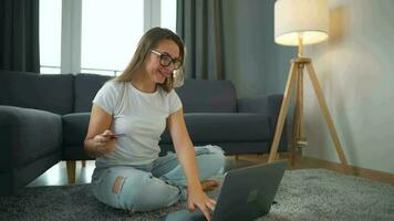 Woman with glasses is sitting on the carpet and makes an online purchase using a credit card and smartphone. Online shopping, lifestyle technology video