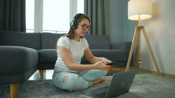 terloops gekleed vrouw met hoofdtelefoons is zittend Aan tapijt met laptop in knus kamer. ze communiceert en werken van een afstand. afgelegen werk buiten de kantoor. video