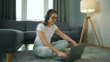 casualmente vestido mulher com fones de ouvido é sentado em tapete com computador portátil dentro acolhedor sala. ela comunica e trabalho remotamente. controlo remoto trabalhos lado de fora a escritório. video