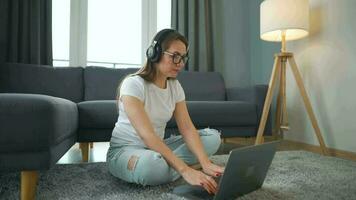 Casually dressed woman with headphones is sitting on carpet with laptop in cozy room. She communicates and works remotely. Remote work outside the office. video