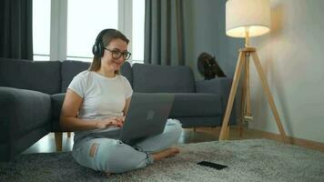 terloops gekleed vrouw met hoofdtelefoons is zittend Aan tapijt met laptop in knus kamer. ze communiceert en werken van een afstand. afgelegen werk buiten de kantoor. video