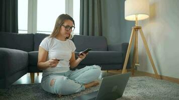 Woman with glasses is sitting on the carpet and makes an online purchase using a credit card and smartphone. Online shopping, lifestyle technology video