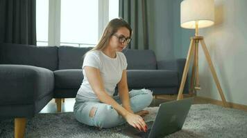 terloops gekleed vrouw is zittend Aan tapijt met laptop in knus kamer. ze communiceert en werken van een afstand. afgelegen werk buiten de kantoor. video