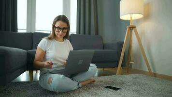 Woman with glasses is sitting on the carpet and makes an online purchase using a credit card and smartphone. Online shopping, lifestyle technology video