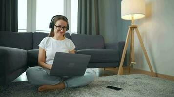 terloops gekleed vrouw met hoofdtelefoons is zittend Aan tapijt met laptop in knus kamer. ze communiceert en werken van een afstand. afgelegen werk buiten de kantoor. video