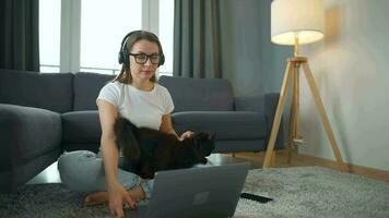 Casually dressed woman with headphones is sitting on carpet with laptop in cozy room. She communicates and works remotely. Remote work outside the office. video