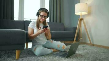 Casually dressed woman with headphones is sitting on carpet with laptop in cozy room. She communicates and works remotely. Remote work outside the office. video