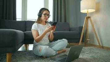 por casualidad vestido mujer con auriculares es sentado en alfombra con ordenador portátil y teléfono inteligente en acogedor habitación. ella comunica y trabajos remotamente remoto trabajo fuera de el oficina. video