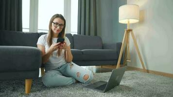 por casualidad vestido mujer con lentes sentado en alfombra con ordenador portátil y teléfono inteligente y trabajando en acogedor habitación. ella comunica y trabajos remotamente remoto trabajo fuera de el oficina. video