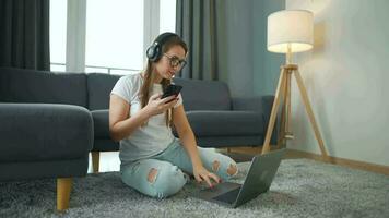 casualmente vestido mulher com fones de ouvido é sentado em tapete com computador portátil e Smartphone dentro acolhedor sala. ela comunica e trabalho remotamente. controlo remoto trabalhos lado de fora a escritório. video