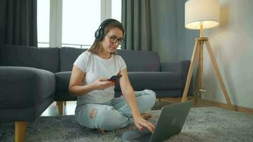 casualmente vestido mulher com fones de ouvido é sentado em tapete com computador portátil e Smartphone dentro acolhedor sala. ela comunica e trabalho remotamente. controlo remoto trabalhos lado de fora a escritório. video