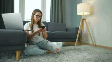 por casualidad vestido mujer con lentes sentado en alfombra con ordenador portátil y teléfono inteligente y trabajando en acogedor habitación. ella comunica y trabajos remotamente remoto trabajo fuera de el oficina. video