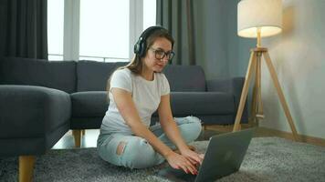 terloops gekleed vrouw met hoofdtelefoons is zittend Aan tapijt met laptop in knus kamer. ze communiceert en werken van een afstand. afgelegen werk buiten de kantoor. video