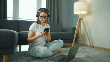 mine de rien habillé femme avec écouteurs est séance sur tapis avec portable et téléphone intelligent dans confortable chambre. elle communique et travaux à distance. éloigné travail à l'extérieur le bureau. video