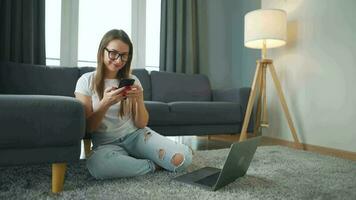 casualmente vestito donna con bicchieri seduta su tappeto con il computer portatile e smartphone e Lavorando nel accogliente camera. lei comunica e lavori a distanza. a distanza opera al di fuori il ufficio. video