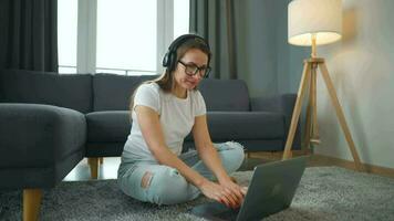 mine de rien habillé femme avec écouteurs est séance sur tapis avec portable dans confortable chambre. elle communique et travaux à distance. éloigné travail à l'extérieur le bureau. video