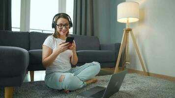 casualmente vestido mulher com fones de ouvido é sentado em tapete com computador portátil e Smartphone dentro acolhedor sala. ela comunica e trabalho remotamente. controlo remoto trabalhos lado de fora a escritório. video