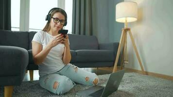 mine de rien habillé femme avec écouteurs est séance sur tapis avec portable et téléphone intelligent dans confortable chambre. elle communique et travaux à distance. éloigné travail à l'extérieur le bureau. video