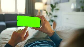 Woman at home lying on a bed and using smartphone with green mock-up screen in horizontal mode. Girl browsing Internet video