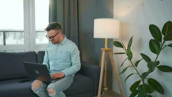 casual vestido homem com óculos sentado em a sofá e usando computador portátil para faço uma vídeo ligar a partir de casa dentro uma moderno apartamento video