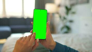 Woman at home lying on a bed and using smartphone with green mock-up screen in vertical mode. video