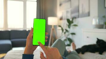 Woman at home lying on a bed and using smartphone with green mock-up screen in vertical mode. video