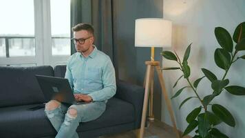 Casual dressed man with glasses sitting on the couch and using laptop to make a video call from home in a modern apartment