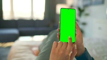 Woman at home lying on a bed and using smartphone with green mock-up screen in vertical mode. video