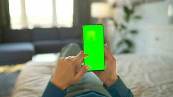 Woman at home lying on a bed and using smartphone with green mock-up screen in vertical mode. video