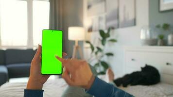 Woman at home lying on a bed and using smartphone with green mock-up screen in vertical mode. video