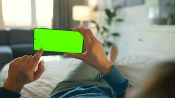 Woman at home lying on a bed and using smartphone with green mock-up screen in horizontal mode. Girl browsing Internet video
