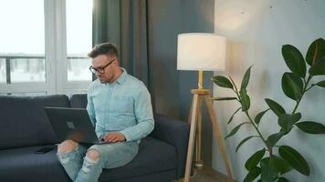 Casual dressed man with glasses sitting on the couch and using laptop to make a video call from home in a modern apartment