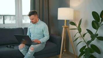 Man with glasses sitting on the sofa with laptop and working in cozy room. Remote work outside the office video