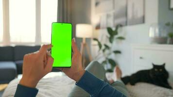 Woman at home lying on a bed and using smartphone with green mock-up screen in vertical mode. video