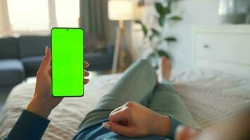 Woman at home lying on a bed and using smartphone with green mock-up screen in vertical mode. video