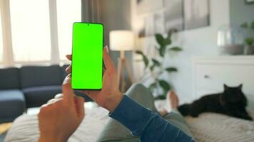 Woman at home lying on a bed and using smartphone with green mock-up screen in vertical mode. video