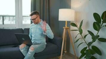 Casual dressed man with glasses sitting on the couch and using laptop to make a video call from home in a modern apartment